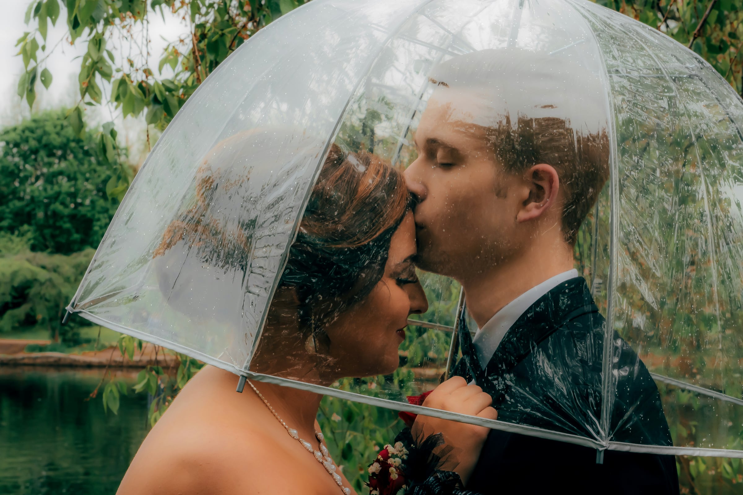 Couple sharing a kiss under a clear umbrella on a rainy day, bride in strapless dress, groom in dark suit