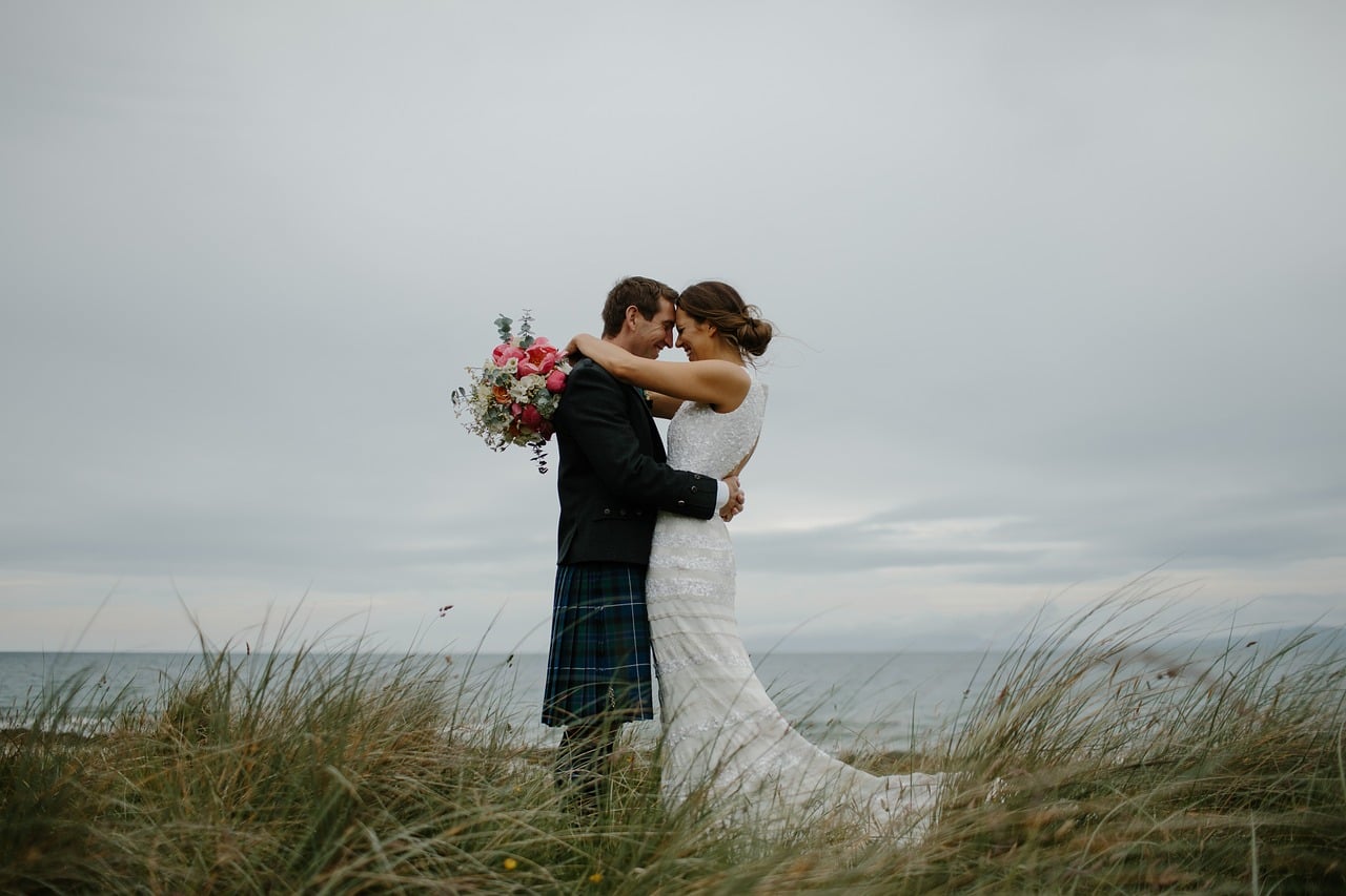 Bride and groom embracing on coastal dunes, groom wearing traditional Scottish kilt, bride holding pink floral bouquet