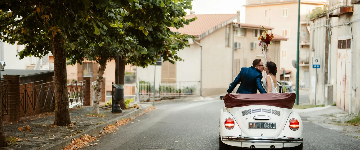 Newlywed couple kissing in white vintage Volkswagen Beetle convertible driving through Italian street, bride holding bouquet