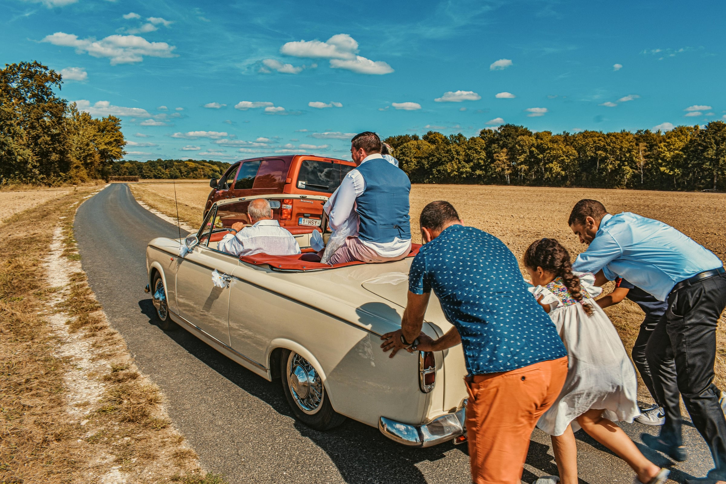 People pushing broken down vintage cream convertible on rural road while couple sits inside, red van ahead
