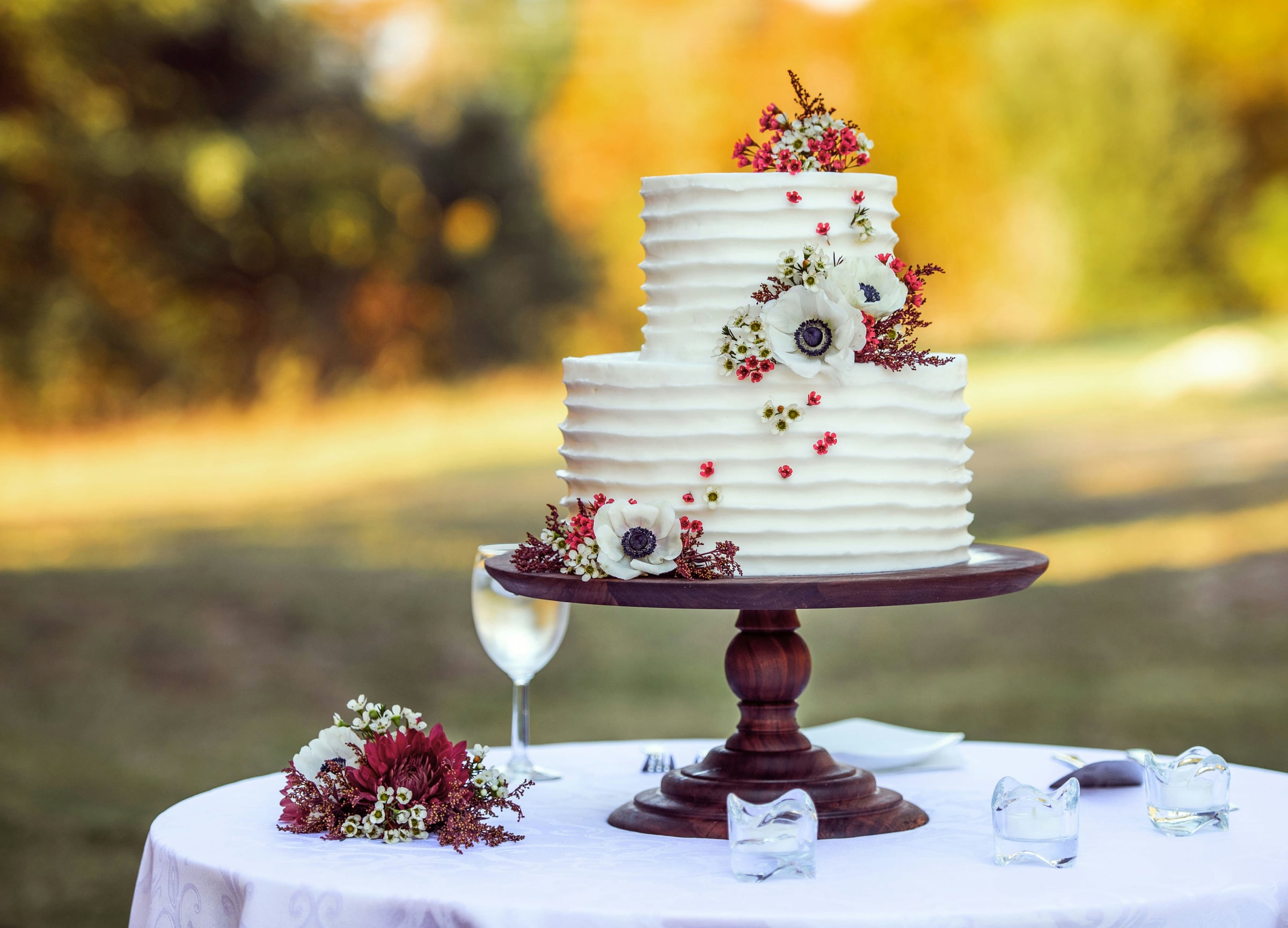 Two-tier white wedding cake with horizontal frosting texture, decorated with white anemones and red flowers on wooden stand,