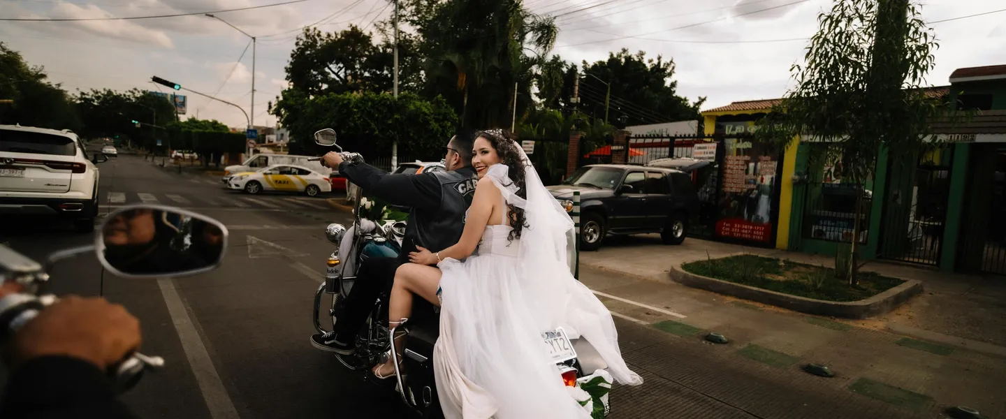 Bride in white wedding dress riding on back of motorcycle with groom on suburban street, her veil and dress flowing behind
