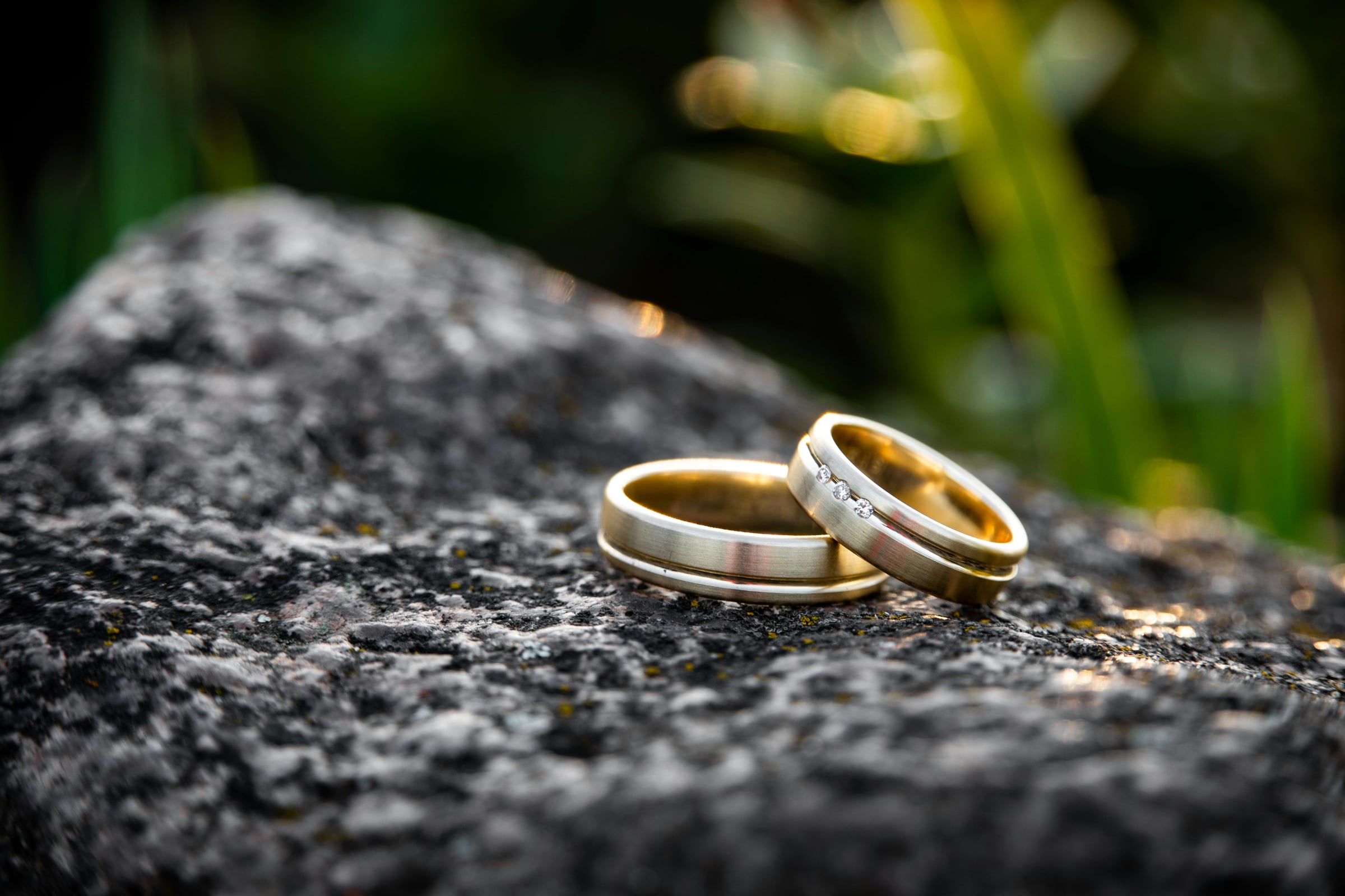 Two gold wedding bands with brushed silver finish resting on a dark granite rock, one featuring three small diamonds
