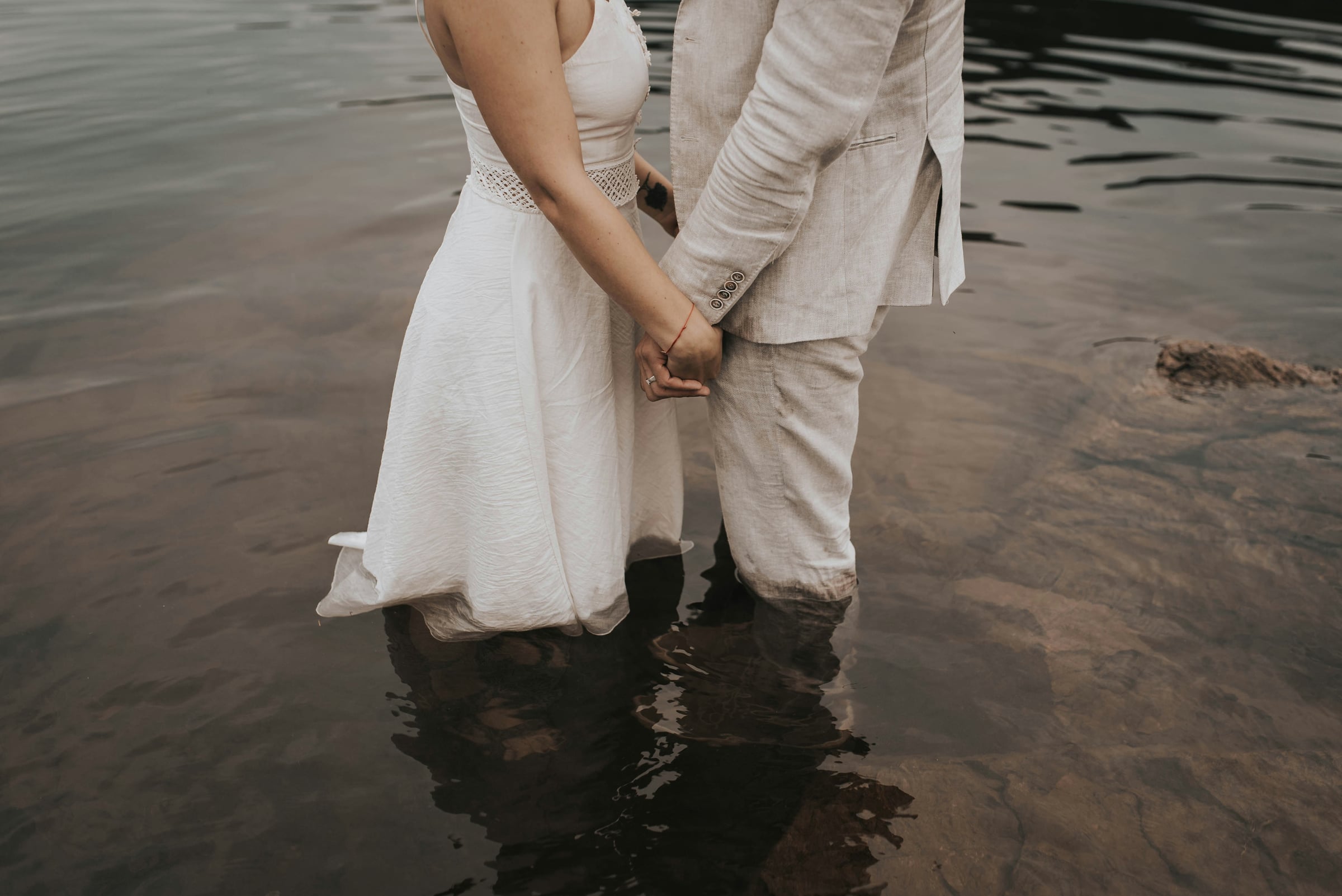 Couple in wedding attire holding hands while standing ankle-deep in calm water, woman in white dress, man in beige linen suit